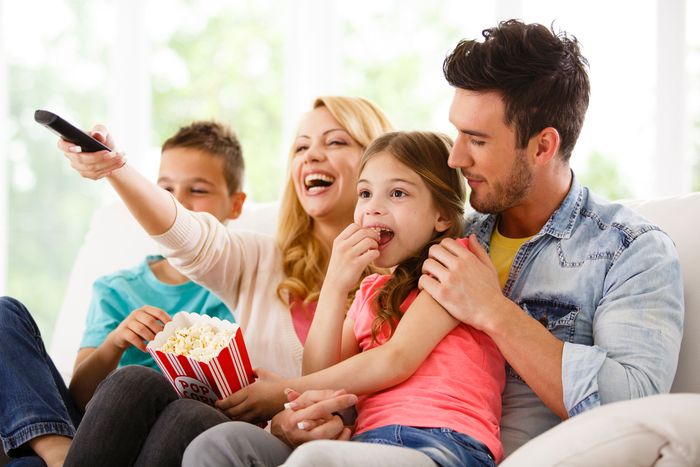 A young mother and father sit on the couch with their young son and daughter. THe mother holds out a TV remote, and the daughter hold a container of popcorn.
